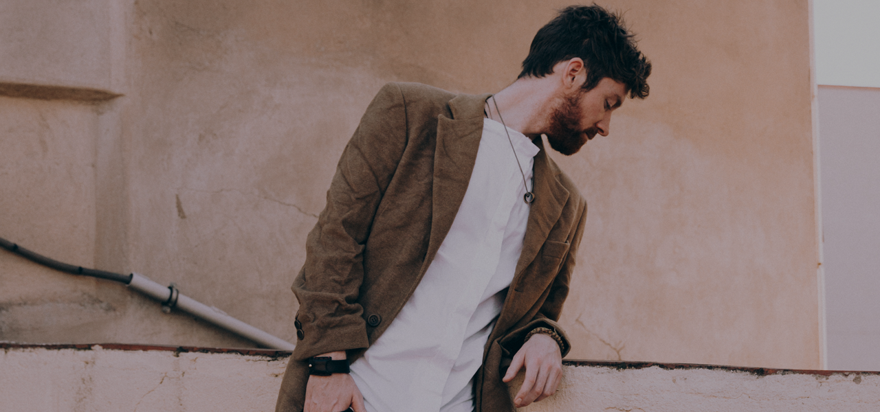 Man in a brown jacket and white shirt standing against a beige wall.